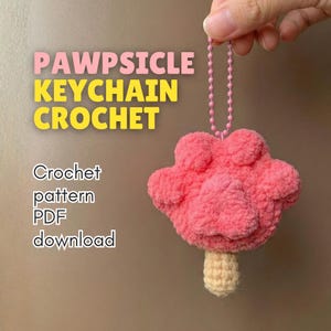 Pode incluir: Um chaveiro rosa feito à mão em crochê em forma de pata, semelhante a um picolé, com um palito bege e uma corrente rosa. O texto "PAWPSICLE KEYCHAIN CROCHET" é exibido acima do chaveiro, com "Crochet pattern PDF download" abaixo.