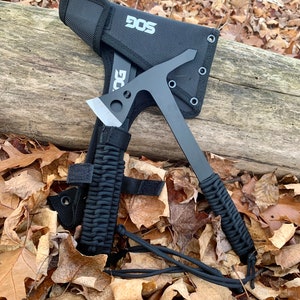May include: A black SOG tomahawk with a black nylon sheath and a paracord handle. The tomahawk is resting on a log with brown leaves scattered around it.