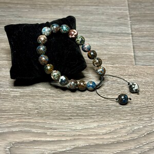 May include: A blue, brown, and white beaded bracelet with a black cord and a tassel. The bracelet is on a black velvet cushion.