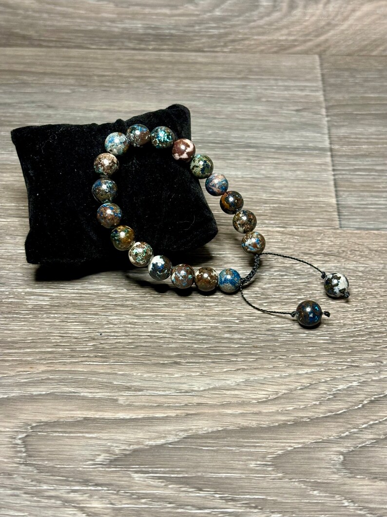 May include: A blue, brown, and white beaded bracelet with a black cord and a tassel. The bracelet is on a black velvet cushion.