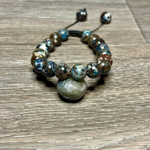 May include: A blue, brown, and white beaded bracelet with a large, round, gray stone charm. The bracelet is made with a black cord and has a sliding knot for adjusting the size.