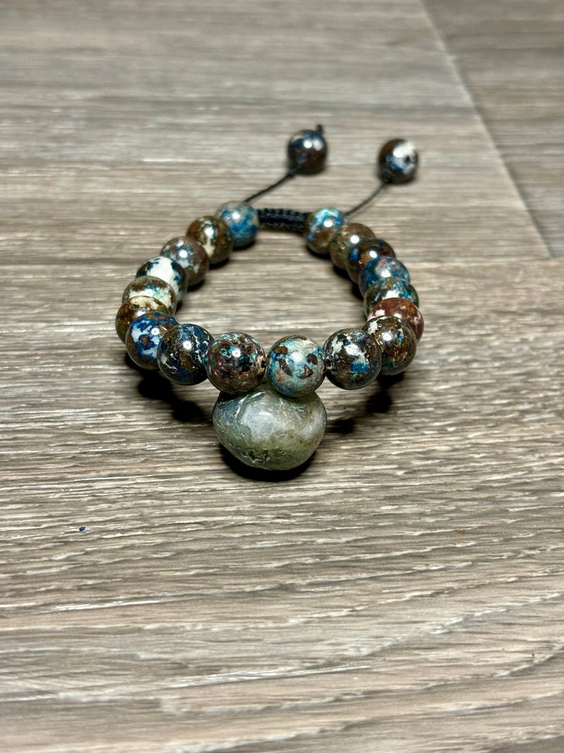 May include: A blue, brown, and white beaded bracelet with a large, round, gray stone charm. The bracelet is made with a black cord and has a sliding knot for adjusting the size.
