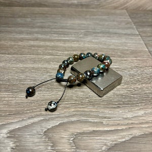 May include: A beaded bracelet with a dark blue, green, and brown color scheme. The bracelet has a black cord with two black beads at the ends. The bracelet is lying on a silver lighter.