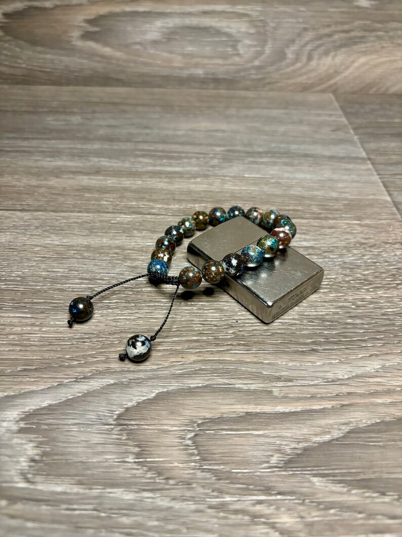May include: A beaded bracelet with a dark blue, green, and brown color scheme. The bracelet has a black cord with two black beads at the ends. The bracelet is lying on a silver lighter.