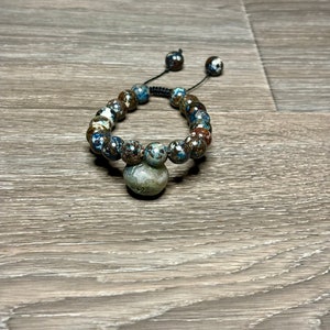 May include: A brown and blue beaded bracelet with a dark gray stone charm. The bracelet is made with a black cord and has a sliding knot for adjusting the size.