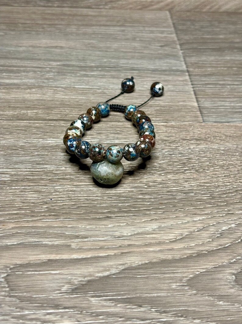 May include: A brown and blue beaded bracelet with a dark gray stone charm. The bracelet is made with a black cord and has a sliding knot for adjusting the size.