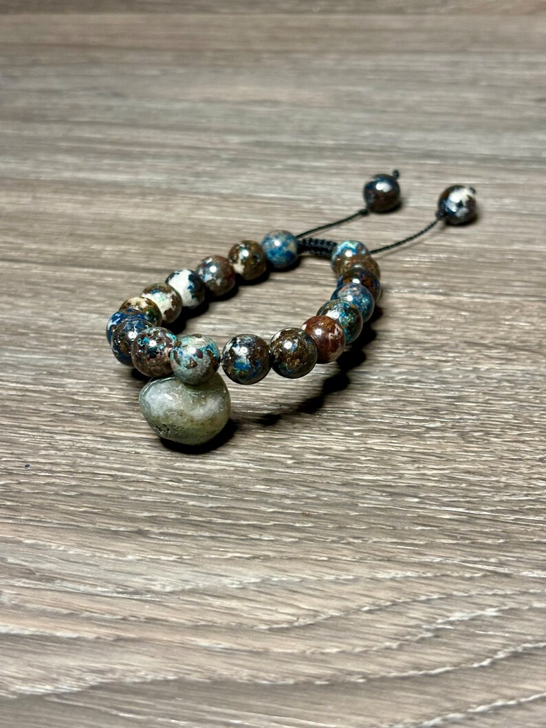 May include: A blue and brown beaded bracelet with a large, round, green and white bead as a focal point. The bracelet is tied with a black cord.