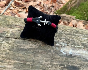 Puede incluir: Un brazalete trenzado negro y rojo con un dije de ancla de plata. El brazalete se muestra en un cojín de terciopelo negro.