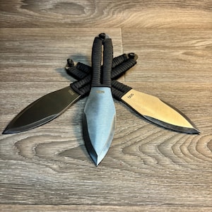 Puede incluir: Tres cuchillos arrojadizos con mangos negros y hojas plateadas dispuestos en forma de estrella. Las hojas tienen la palabra "SOG" impresa. Los mangos están envueltos en un cordón negro.