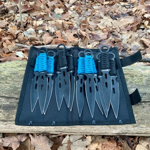 May include: A set of black throwing knives with silver edges and blue and black wrapped handles, displayed in a black carrying case. The knives are arranged in a row, with the blades pointing upwards. The background features fallen leaves.