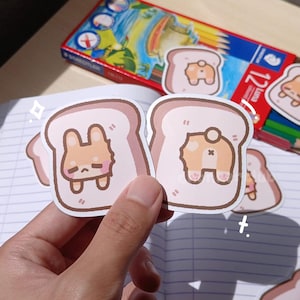May include: Two brown and white cartoon bunny stickers on a white background. The bunnies are inside of a cartoon slice of bread. The stickers are on a notebook with lined paper.