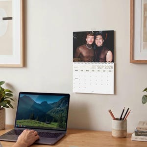 Op de afbeelding: Een wandkalender voor september 2026 met een foto van twee personen in donkere mouwloze tops. De kalender heeft een witte achtergrond met zwarte tekst en een raster voor de dagen van de maand, opgehangen aan een metalen spiraalbinding.