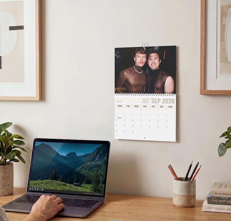 Op de afbeelding: Een wandkalender voor september 2026 met een foto van twee personen in donkere mouwloze tops. De kalender heeft een witte achtergrond met zwarte tekst en een raster voor de dagen van de maand, opgehangen aan een metalen spiraalbinding.