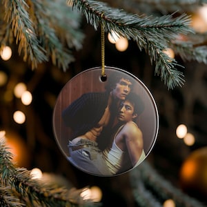 May include: A round ceramic ornament featuring a printed image of two individuals in an intimate embrace. One person wears a dark plaid shirt and grey trousers, while the other wears a white tank top. The ornament hangs from a gold-colored string.