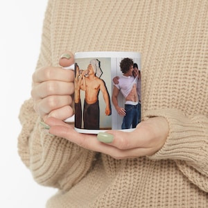 Pode incluir: Caneca de cerâmica branca com duas imagens. Uma mostra uma pessoa bebendo, enrolada em uma toalha. A outra mostra duas pessoas se abraçando. A caneca é segurada por uma pessoa que usa um suéter bege.