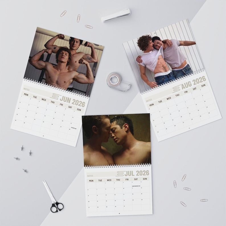 Op de afbeelding: Drie wandkalenderpagina's voor juni, juli en augustus 2026. Elke pagina bevat een andere foto van twee mannen. De juni-pagina toont twee mannen met ontbloot bovenlijf die poseren. Juli toont twee mannen dicht bij elkaar. Augustus toont twee mannen in witte shirts.