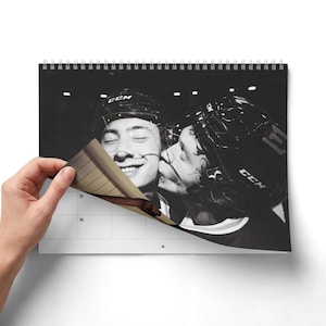 Op de afbeelding: Een wandkalender met spiraalbinding en een zwart-witfoto van twee ijshockeyspelers met CCM-helmen. De ene speler kust de andere op de wang. De kalenderpagina toont een raster met de cijfers 23 en 30 duidelijk zichtbaar.