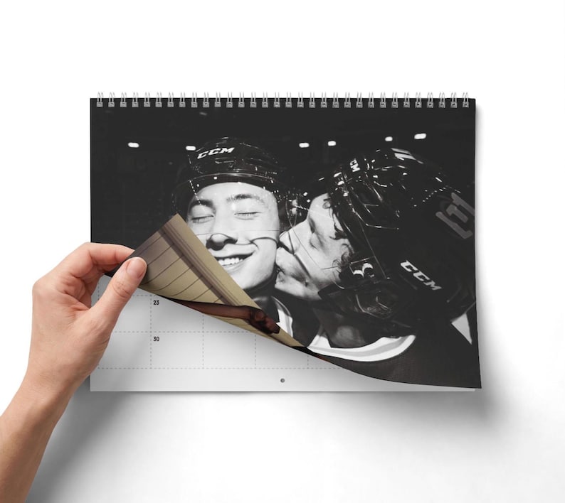 Op de afbeelding: Een wandkalender met spiraalbinding en een zwart-witfoto van twee ijshockeyspelers met CCM-helmen. De ene speler kust de andere op de wang. De kalenderpagina toont een raster met de cijfers 23 en 30 duidelijk zichtbaar.