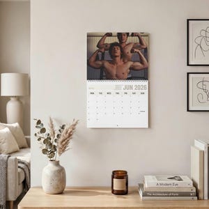 Op de afbeelding: Een wandkalender voor juni 2026 met een foto van twee personen met ontbloot bovenlijf die hun armspieren aanspannen. De kalender heeft een witte achtergrond met zwarte tekst en een raster voor de dagen, gebonden met een zwarte spiraal.