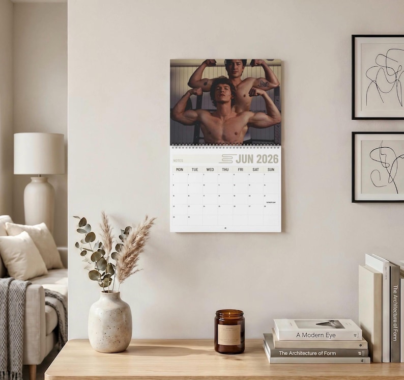 Op de afbeelding: Een wandkalender voor juni 2026 met een foto van twee personen met ontbloot bovenlijf die hun armspieren aanspannen. De kalender heeft een witte achtergrond met zwarte tekst en een raster voor de dagen, gebonden met een zwarte spiraal.