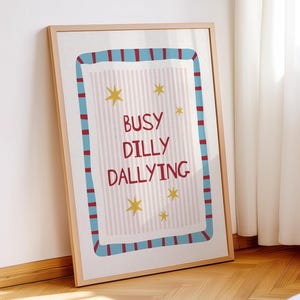 May include: A framed wall art print featuring the text BUSY DILLY DALLYING in red letters. The background has light pink vertical stripes and yellow star illustrations. The border is a thick blue frame with red vertical stripes.