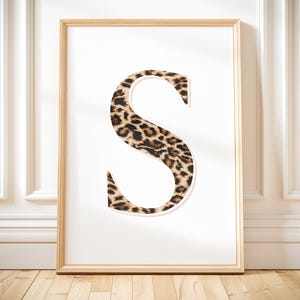 Può includere: Una stampa incorniciata con la lettera "S" in un design audace. La lettera è riempita con un motivo leopardato nei toni del marrone e del nero. L'opera d'arte è ambientata su uno sfondo bianco e incorniciata in legno chiaro.