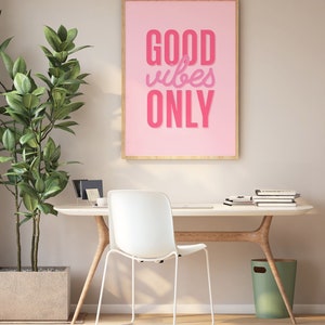May include: A pink framed poster with the text "GOOD vibes ONLY" in pink lettering on a pink background. The poster is hanging on a wall above a white desk with a white chair and a green trash can.