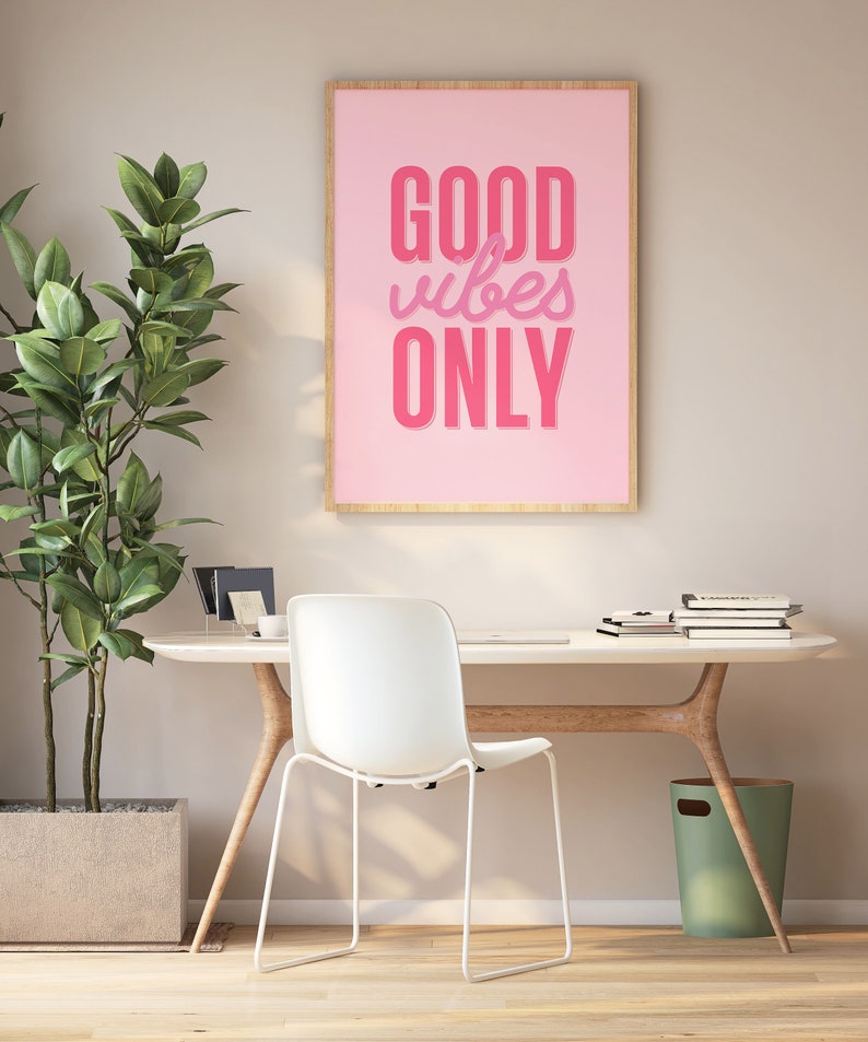 May include: A pink framed poster with the text "GOOD vibes ONLY" in pink lettering on a pink background. The poster is hanging on a wall above a white desk with a white chair and a green trash can.