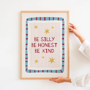 Op de afbeelding: Een rechthoekige wandkunstprint in een lichthouten lijst met de tekst BE SILLY BE HONEST BE KIND in donkerrode letters. De achtergrond heeft lichtroze verticale strepen, gele sterren en een blauwe rand met donkerrode verticale streepjes.