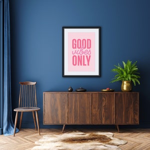 May include: A framed print with a pink background and the text "GOOD vibes ONLY" in pink lettering. The print is hanging on a dark blue wall above a wooden cabinet with a light brown rug in front of it.