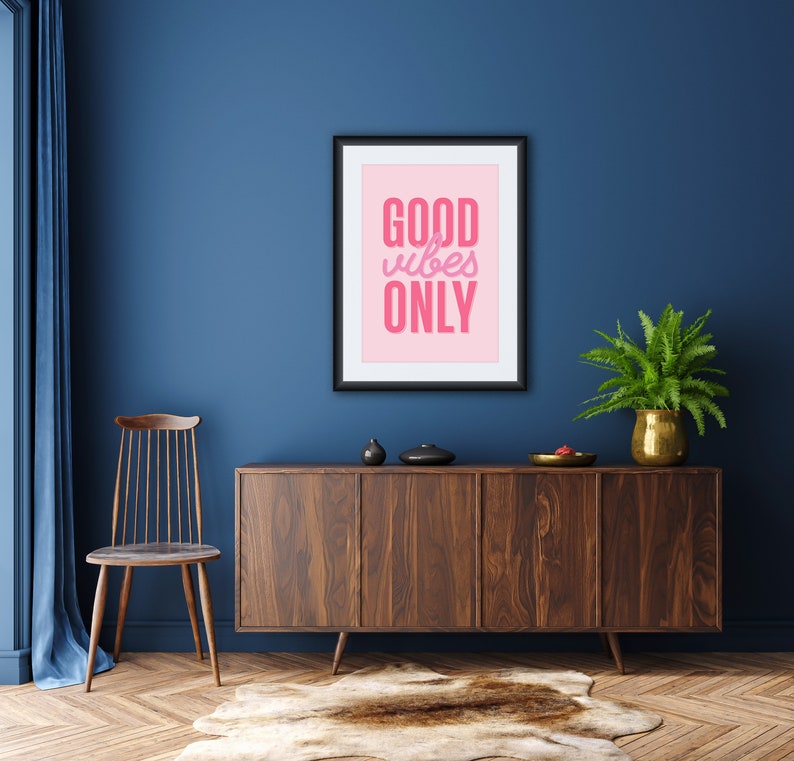 May include: A framed print with a pink background and the text "GOOD vibes ONLY" in pink lettering. The print is hanging on a dark blue wall above a wooden cabinet with a light brown rug in front of it.