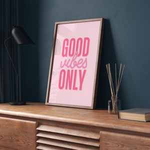 May include: A framed pink poster with the text "GOOD vibes ONLY" in a pink and white font. The poster is hanging on a dark blue wall above a wooden cabinet.