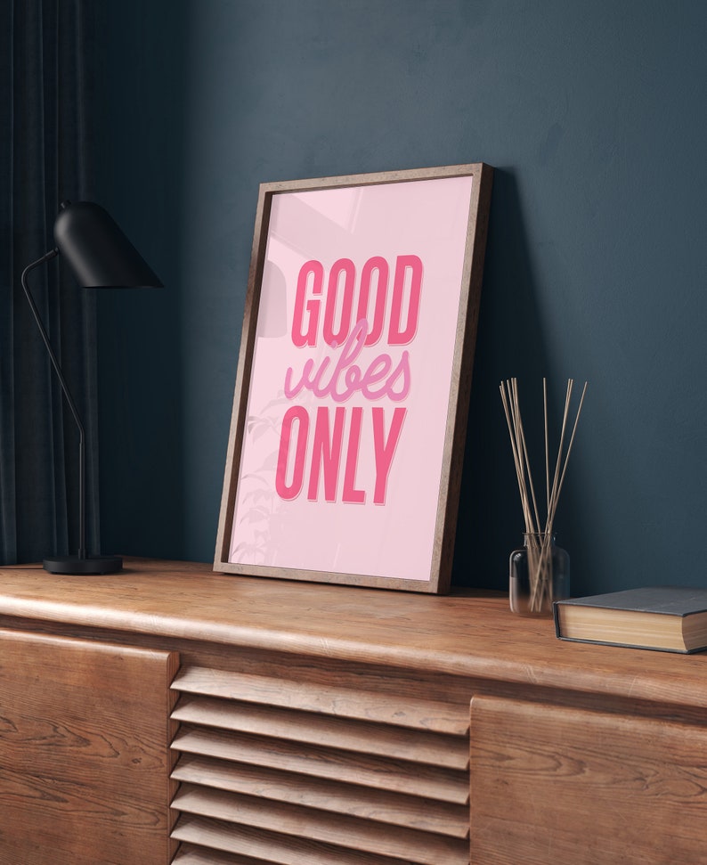 May include: A framed pink poster with the text "GOOD vibes ONLY" in a pink and white font. The poster is hanging on a dark blue wall above a wooden cabinet.