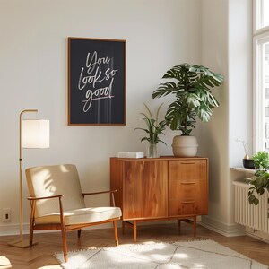 May include: A mid-century modern living room scene featuring a wooden cabinet, a beige armchair, and a floor lamp with a white lampshade. A framed artwork with the text "You look so good" hangs on the wall. A large potted plant adds a touch of nature.
