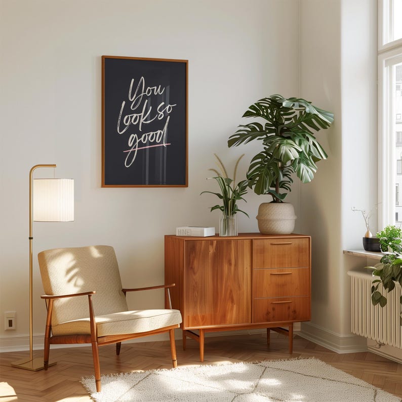 May include: A mid-century modern living room scene featuring a wooden cabinet, a beige armchair, and a floor lamp with a white lampshade. A framed artwork with the text "You look so good" hangs on the wall. A large potted plant adds a touch of nature.