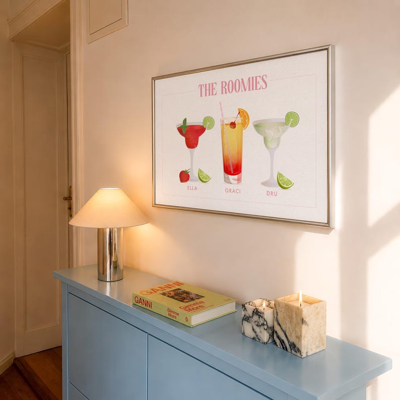 May include: A framed horizontal wall art print titled THE ROOMIES featuring three illustrated cocktails with garnishes. The drinks are labeled ELLA, GRACI, and DRU in pink text. The artwork displays a red drink, an orange drink, and a light green drink.