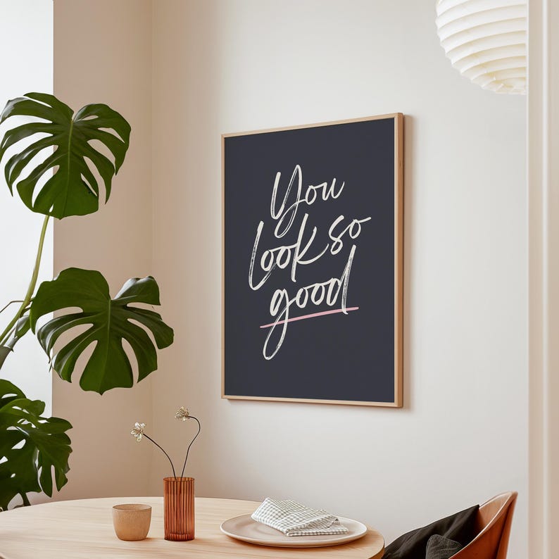 May include: A framed art print with the words "You look so good" in white script on a dark blue background. The print has a light wood frame and a pink line underneath the word "good". The print is hanging on a white wall.