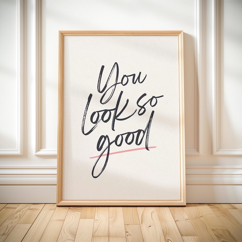 May include: A framed print with the words "You look so good" in black script. The words are on a white background with a thin pink line underneath "good". The frame is light brown, and the print is leaning against a white wall on a wooden floor.