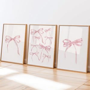 Può includere: Tre stampe artistiche incorniciate con illustrazioni ad acquerello di fiocchi rosa. I fiocchi variano per dimensioni e stile, con lunghi nastri fluenti. Le cornici sono di colore legno chiaro e lo sfondo è bianco sporco neutro. L'opera d'arte è esposta su un pavimento in legno.