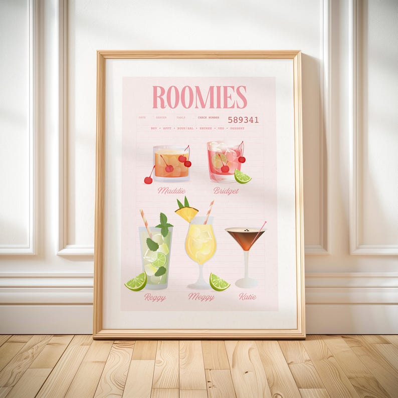 Op de afbeelding: Een ingelijste print met het woord "ROOMIES" in roze, met illustraties van cocktails. De print bevat namen en recepten. Cocktails zijn gegarneerd met kersen, ananas en limoen. De lijst is licht van kleur.