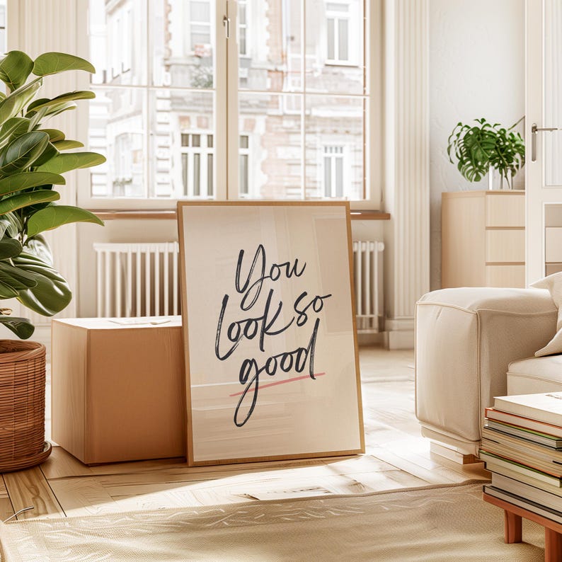 May include: A framed print with the words "You look so good" in black script. The print has a light beige background and a thin wooden frame. The print is leaning against a cardboard box in a well-lit room.