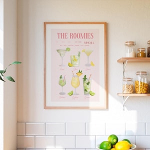 Op de afbeelding: Een ingelijste print met aquarel illustraties van verschillende cocktails, waaronder margaritas en mojito's, op een lichtroze achtergrond. De print heeft de titel "THE ROOMIES" en bevat een reeks cocktailnamen. De print wordt weergegeven op een witte betegelde muur.