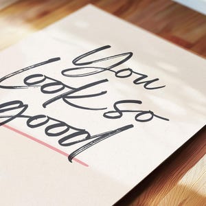 May include: A greeting card with the words "You look so good" in a handwritten, dark gray script. A thin pink line underlines the word "good". The card is on a light beige background, with a wooden surface in the foreground.