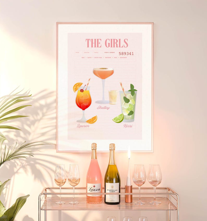 Op de afbeelding: Een ingelijste print getiteld "The Girls" met illustraties van cocktails en namen. Daaronder een bartrol met twee flessen mousserende wijn, een brandende kaars en wijnglazen. De print en de kar staan tegen een lichtroze muur.