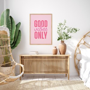 May include: A pink framed poster with the text "GOOD vibes ONLY" in a pink, stylized font. The poster is hanging on a white wall above a wooden cabinet with a woven design. There is a hanging chair and a potted plant in the background.