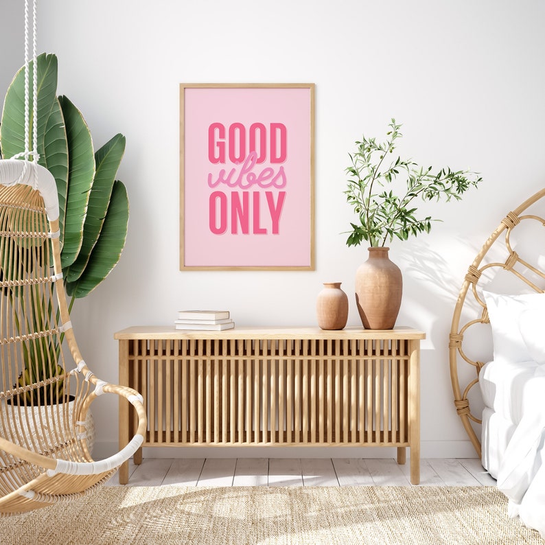 May include: A pink framed poster with the text "GOOD vibes ONLY" in a pink, stylized font. The poster is hanging on a white wall above a wooden cabinet with a woven design. There is a hanging chair and a potted plant in the background.
