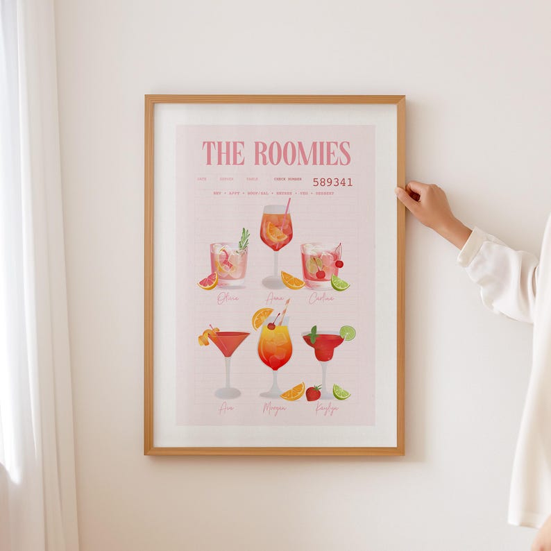 Op de afbeelding: Een ingelijste print getiteld "The Roomies" met illustraties van cocktails. Het kunstwerk bevat zes verschillende cocktailglazen met diverse garneringen zoals sinaasappelschijfjes, kersen en limoenen. De print heeft een lichtroze achtergrond en zit in een houten lijst.