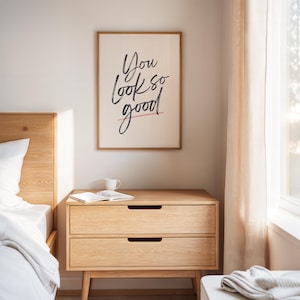 May include: A framed print with the words "You look so good" in black script with a pink underline. The print hangs above a light wood nightstand with two drawers. A book and a white mug sit on top of the nightstand.