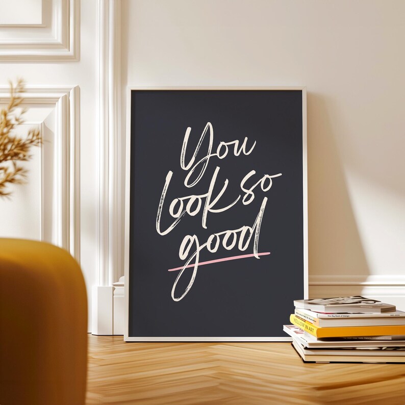 May include: A framed art print with the words "You look so good" in white script on a dark gray background. A thin pink line underlines the word "good". The print is displayed on a wooden floor.