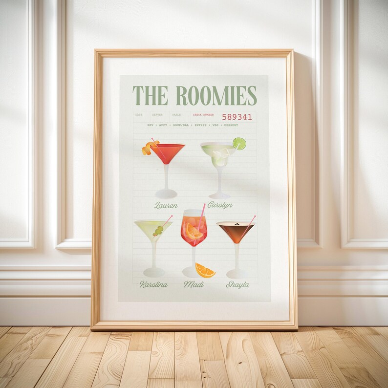 Op de afbeelding: Een ingelijste print met de tekst "THE ROOMIES" en illustraties van zes verschillende cocktails. Elke cocktail is voorzien van een naam. De print zit in een licht houten frame en wordt gepresenteerd tegen een witte muur en houten vloer.